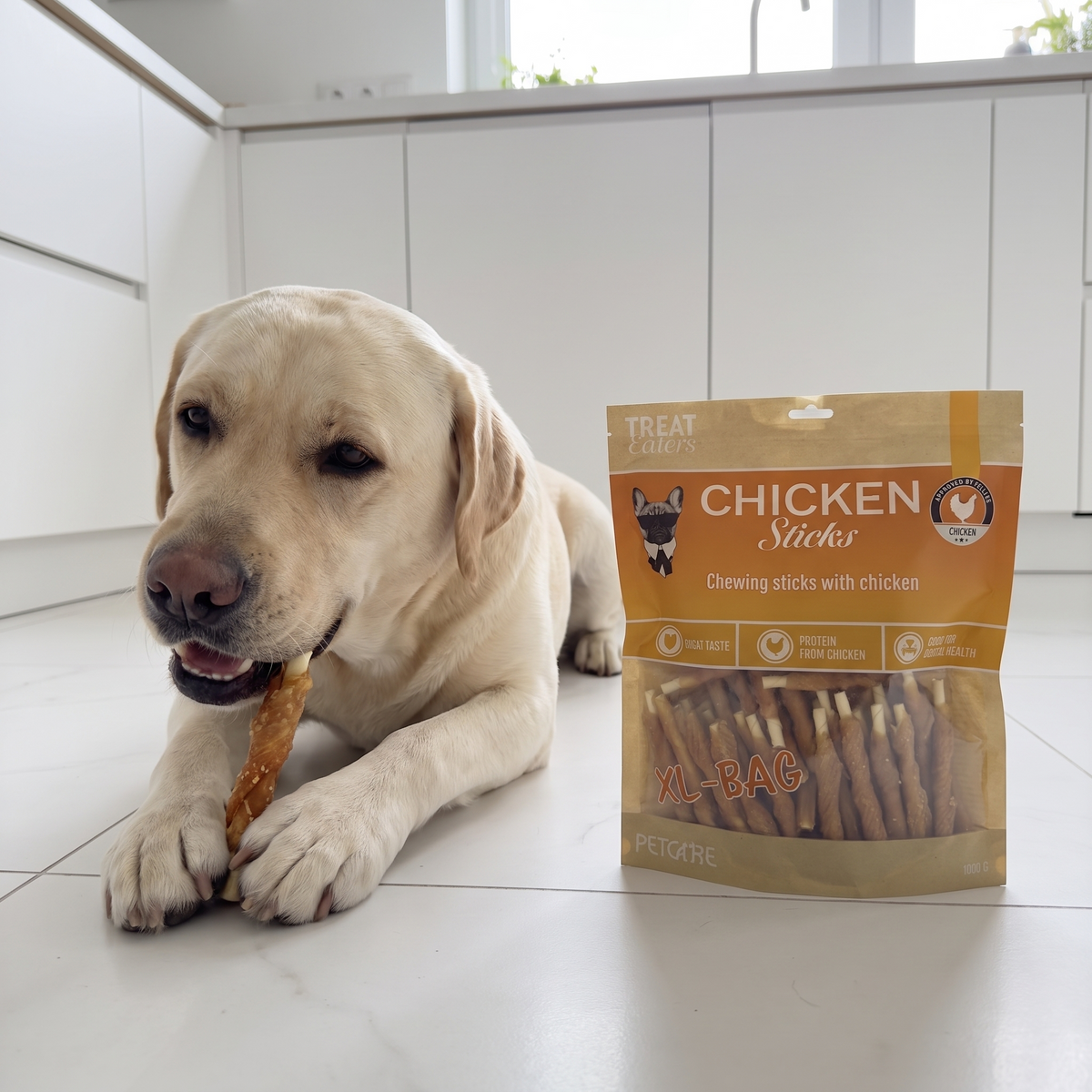 Gul labrador ligger og tygger på en kyllingestick i et lyst køkken, med posen ved siden af