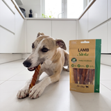 Whippet ligger og tygger på en lametyggestang i et lyst køkken, med posen ved siden af